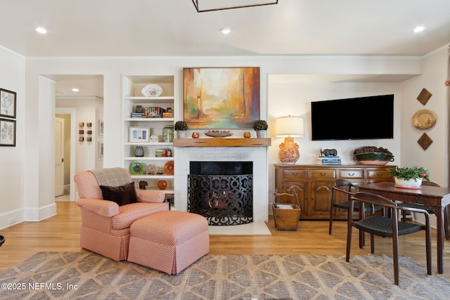 living room featuring built in shelves, ornamental molding, a tiled fireplace, and light wood-type flooring