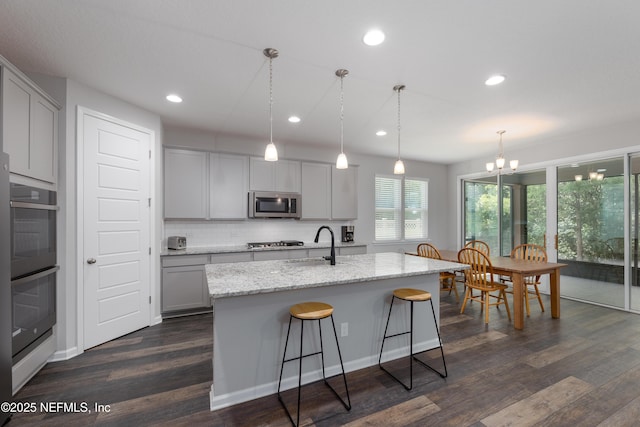 kitchen with stainless steel appliances, tasteful backsplash, light stone countertops, a center island with sink, and decorative light fixtures