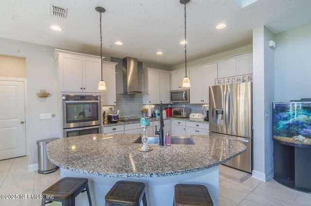 kitchen with a kitchen island with sink, visible vents, appliances with stainless steel finishes, wall chimney exhaust hood, and a kitchen bar