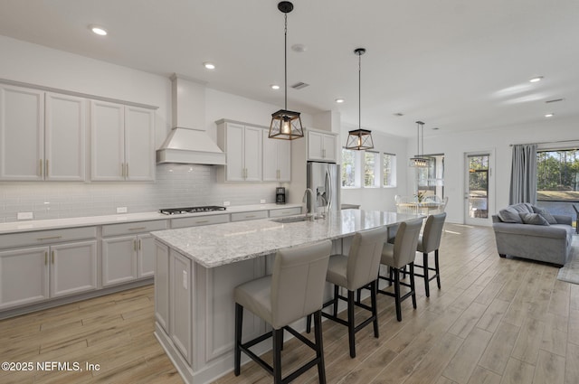 kitchen with hanging light fixtures, gas stovetop, custom range hood, an island with sink, and stainless steel fridge with ice dispenser