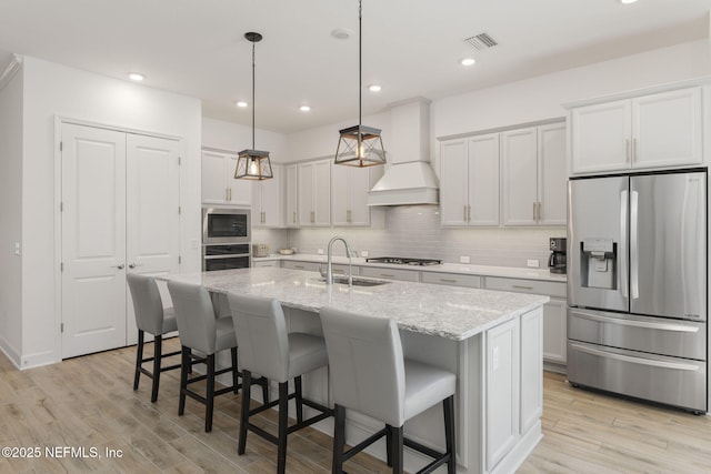 kitchen featuring sink, premium range hood, stainless steel appliances, a center island with sink, and decorative light fixtures