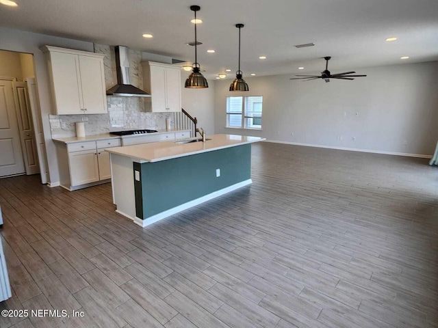 kitchen featuring a center island with sink, white cabinetry, wall chimney range hood, and decorative light fixtures