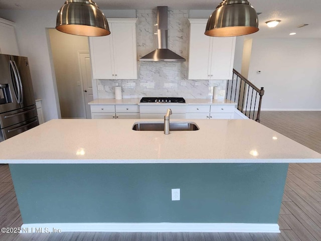 kitchen with sink, wall chimney range hood, light stone counters, stainless steel refrigerator with ice dispenser, and a center island with sink