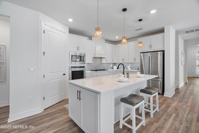 kitchen featuring appliances with stainless steel finishes, a kitchen island with sink, decorative backsplash, white cabinets, and decorative light fixtures