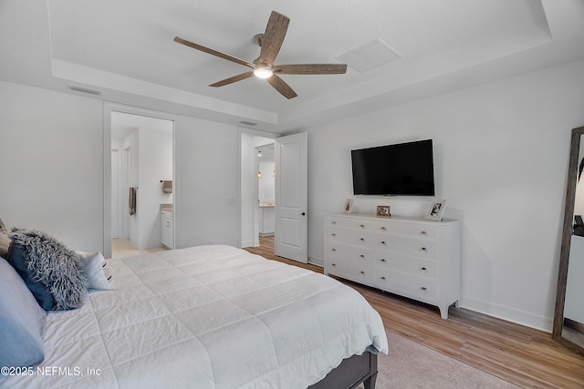 bedroom with ceiling fan, ensuite bath, a raised ceiling, and light hardwood / wood-style flooring