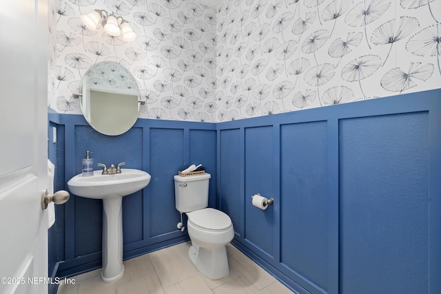 half bath featuring a wainscoted wall, a decorative wall, toilet, a sink, and wallpapered walls