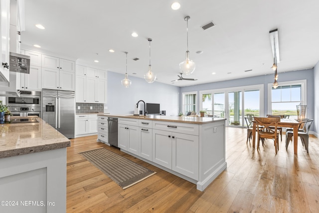 kitchen featuring appliances with stainless steel finishes, decorative light fixtures, white cabinets, light stone counters, and a center island with sink