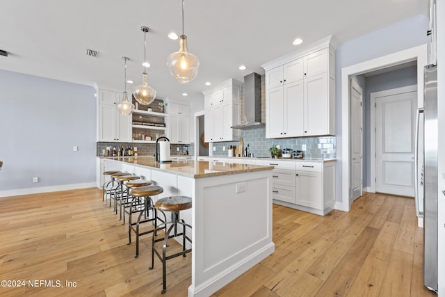 kitchen featuring light stone counters, hanging light fixtures, a center island with sink, wall chimney range hood, and white cabinets