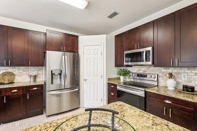 kitchen with a textured ceiling, light tile patterned floors, appliances with stainless steel finishes, light stone countertops, and decorative backsplash