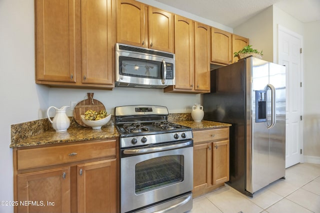 kitchen with dark stone countertops, stainless steel appliances, brown cabinets, and light tile patterned flooring