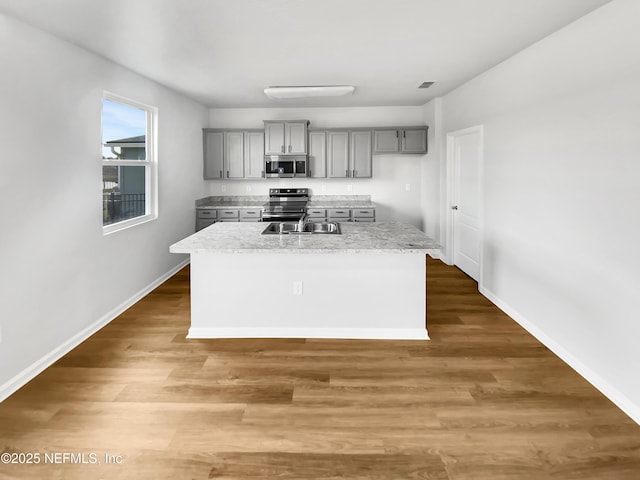 kitchen with light stone countertops, gray cabinets, stainless steel appliances, and a center island with sink