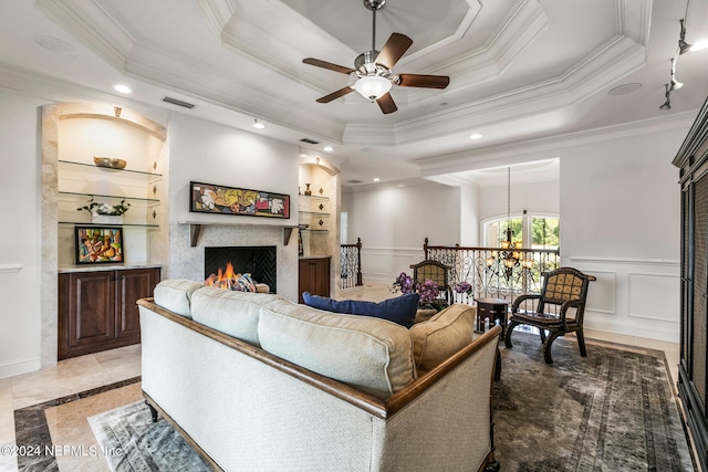 living room with ceiling fan with notable chandelier, built in features, a fireplace, ornamental molding, and a tray ceiling