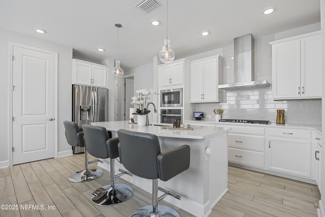 kitchen featuring a center island with sink, wall chimney range hood, white cabinets, and decorative light fixtures