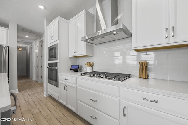 kitchen featuring appliances with stainless steel finishes, white cabinets, wall chimney range hood, light stone countertops, and backsplash