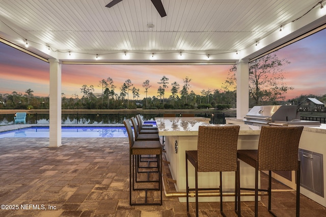 patio terrace at dusk featuring an outdoor bar, ceiling fan, a water view, a grill, and exterior kitchen