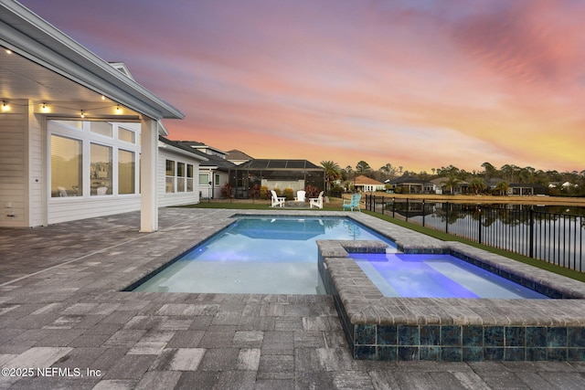 pool at dusk with an in ground hot tub, a water view, and a patio