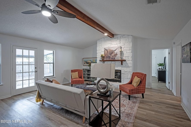 living room with a stone fireplace, lofted ceiling with beams, ceiling fan, a textured ceiling, and light hardwood / wood-style flooring