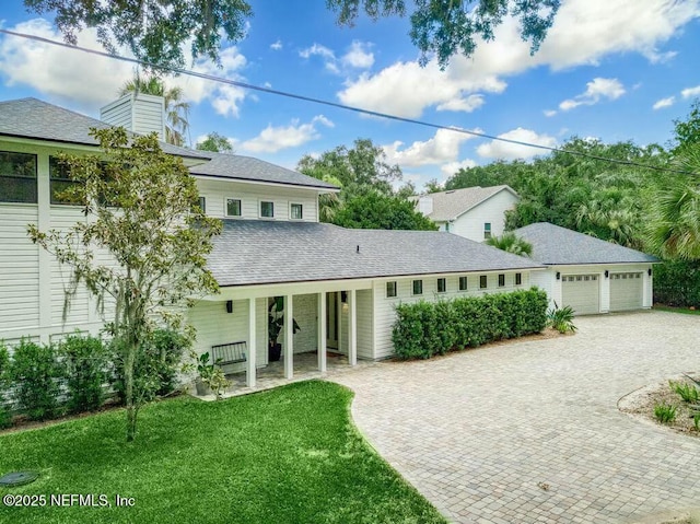 view of front of house with covered porch, roof with shingles, decorative driveway, a front lawn, and a chimney