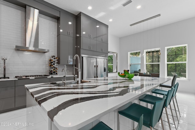 kitchen featuring wall chimney exhaust hood, a breakfast bar, appliances with stainless steel finishes, gray cabinets, and backsplash