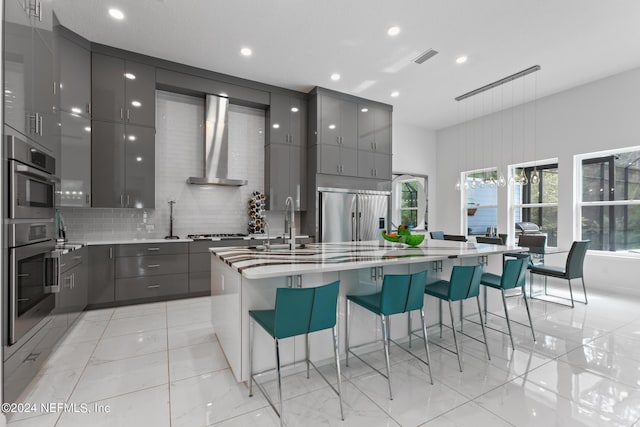 kitchen with stainless steel appliances, an island with sink, a kitchen bar, and wall chimney exhaust hood