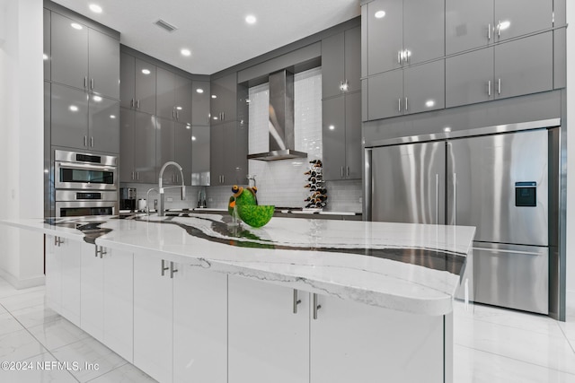 kitchen featuring wall chimney range hood, gray cabinetry, light stone counters, built in fridge, and stainless steel double oven