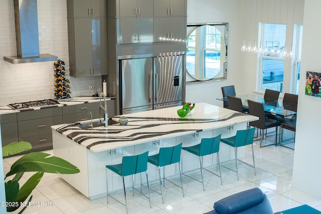 kitchen featuring wall chimney range hood, gray cabinets, stainless steel appliances, a kitchen breakfast bar, and tasteful backsplash