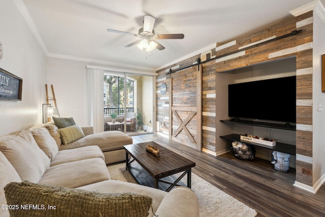 living room with wooden walls, ornamental molding, ceiling fan, a barn door, and dark wood-type flooring