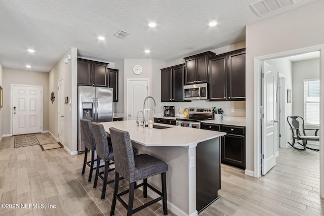 kitchen with light wood finished floors, appliances with stainless steel finishes, a sink, an island with sink, and a kitchen bar