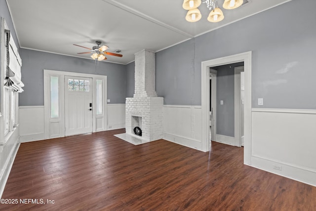 unfurnished living room with ceiling fan with notable chandelier, dark wood-type flooring, a brick fireplace, and crown molding