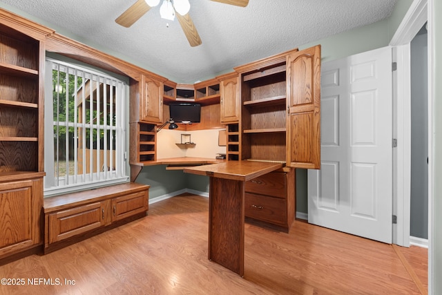 unfurnished office with a textured ceiling, ceiling fan, baseboards, light wood-style floors, and built in desk