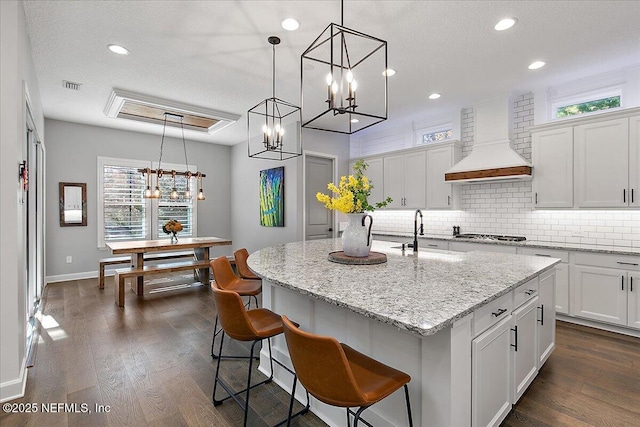 kitchen with premium range hood, sink, white cabinetry, hanging light fixtures, and a kitchen island with sink