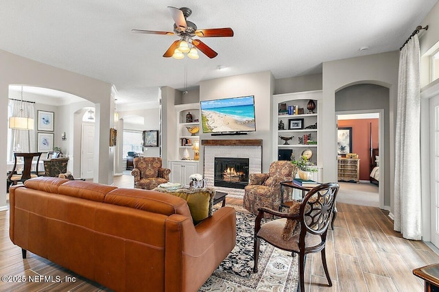 living room featuring built in shelves, ceiling fan, a brick fireplace, a textured ceiling, and light hardwood / wood-style flooring