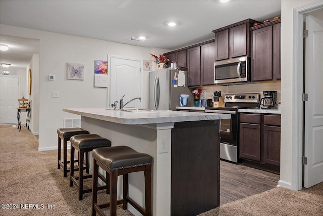 kitchen with a kitchen island with sink, dark brown cabinets, stainless steel appliances, a kitchen bar, and dark carpet