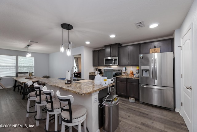 kitchen featuring decorative light fixtures, appliances with stainless steel finishes, a kitchen island with sink, a sink, and light stone countertops