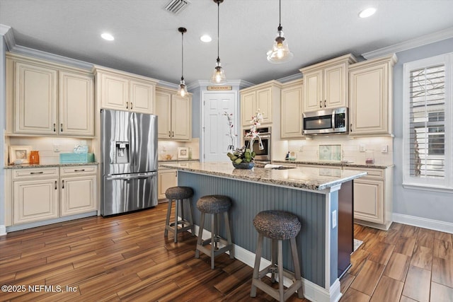 kitchen featuring appliances with stainless steel finishes, hanging light fixtures, light stone counters, a center island with sink, and cream cabinetry