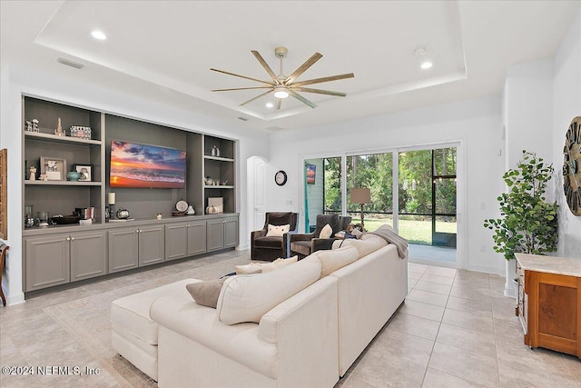 living room featuring light tile patterned flooring, built in shelves, ceiling fan, and a tray ceiling