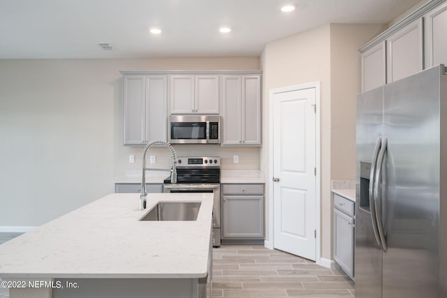 kitchen featuring gray cabinetry, light stone counters, a kitchen island with sink, and appliances with stainless steel finishes