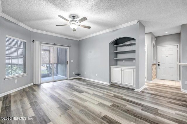 unfurnished living room featuring a textured ceiling, built in shelves, light wood-style flooring, baseboards, and ornamental molding