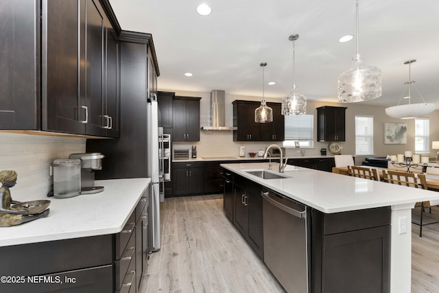 kitchen featuring sink, hanging light fixtures, a kitchen island with sink, stainless steel dishwasher, and wall chimney exhaust hood