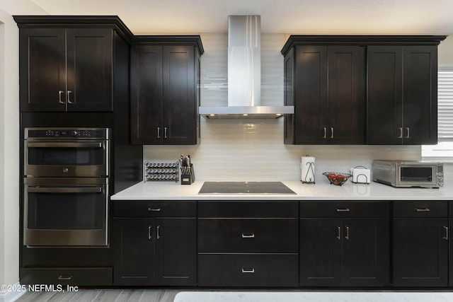 kitchen featuring dark brown cabinetry, black electric cooktop, stainless steel double oven, wall chimney range hood, and backsplash