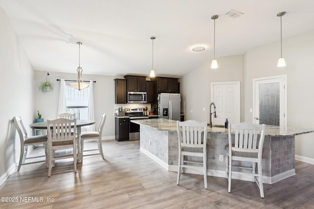 kitchen with dark brown cabinetry, hanging light fixtures, appliances with stainless steel finishes, light stone countertops, and a kitchen island with sink