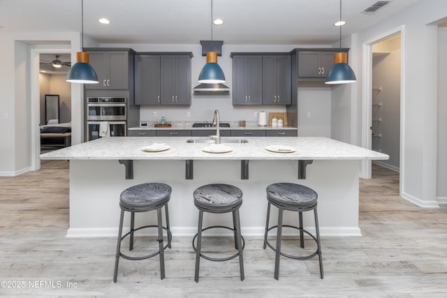 kitchen with light stone counters, a kitchen island with sink, double oven, and hanging light fixtures