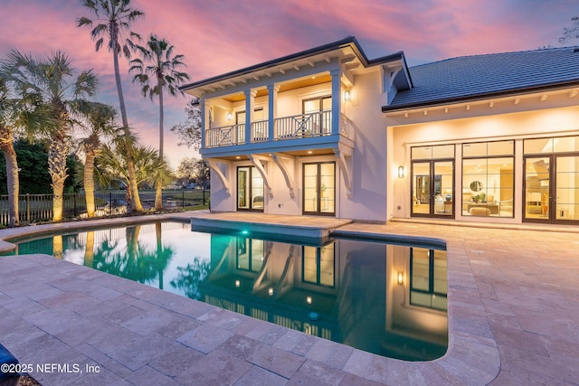 back of property at dusk featuring french doors, stucco siding, a patio area, fence, and a balcony