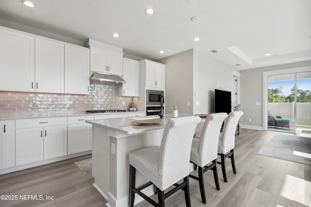 kitchen with stainless steel microwave, tasteful backsplash, light stone countertops, white cabinets, and a center island with sink