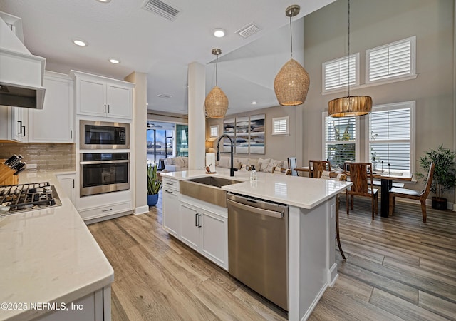 kitchen featuring white cabinetry, hanging light fixtures, stainless steel appliances, light stone counters, and an island with sink