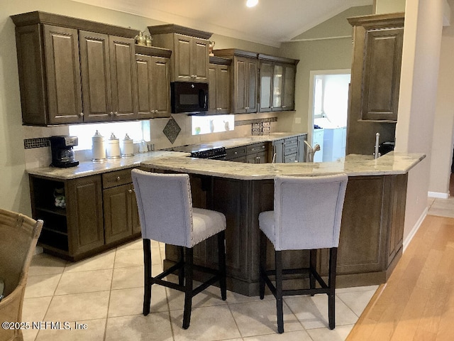 kitchen with a breakfast bar area, vaulted ceiling, light stone countertops, black microwave, and a peninsula