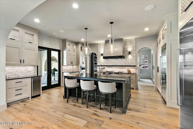 kitchen with premium range hood, decorative light fixtures, a center island with sink, light wood-type flooring, and decorative backsplash