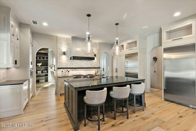 kitchen with white cabinetry, an island with sink, built in refrigerator, hanging light fixtures, and wall chimney range hood