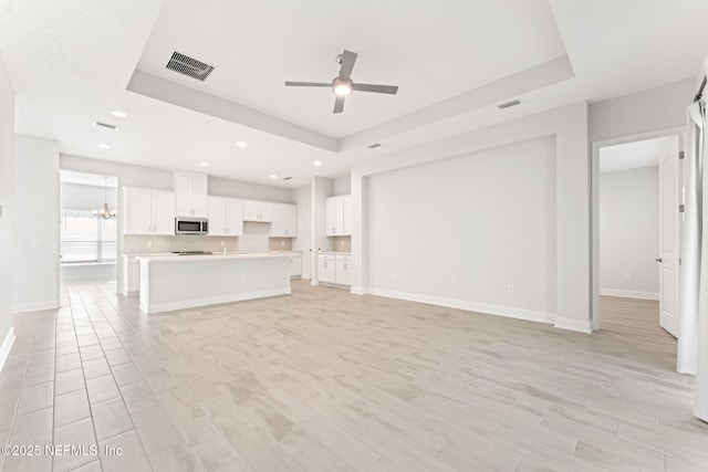 unfurnished living room featuring a raised ceiling, ceiling fan with notable chandelier, and light wood-type flooring