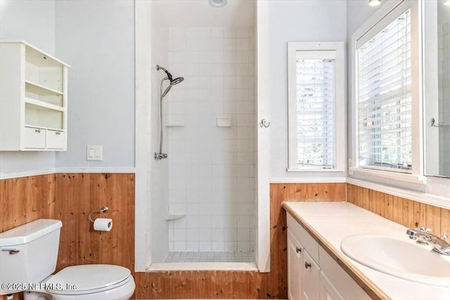 bathroom featuring toilet, a wainscoted wall, wood walls, vanity, and tiled shower